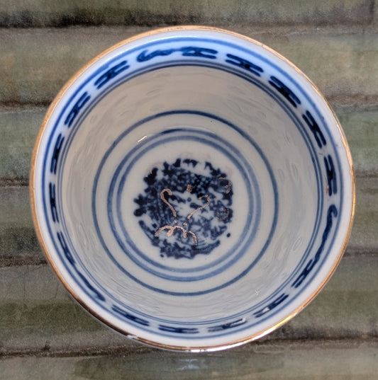 A top-down view of a ceramic cup with a white glaze, decorated with blue concentric circles, a blue geometric border, and a small blue floral motif in the center. The cup has a thin gold rim and is resting on a textured green surface.