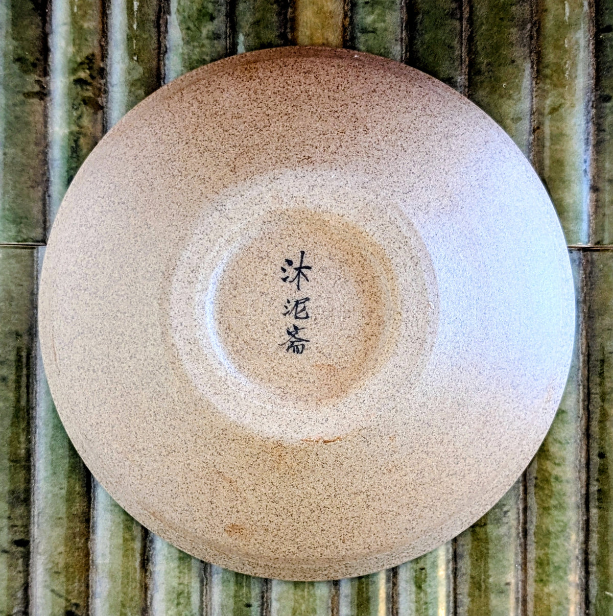 A round ceramic bowl photographed from above on a green bamboo surface. The bowl has a speckled warm beige glaze with a darker glaze ring around the center and vertical black calligraphy characters printed inside.