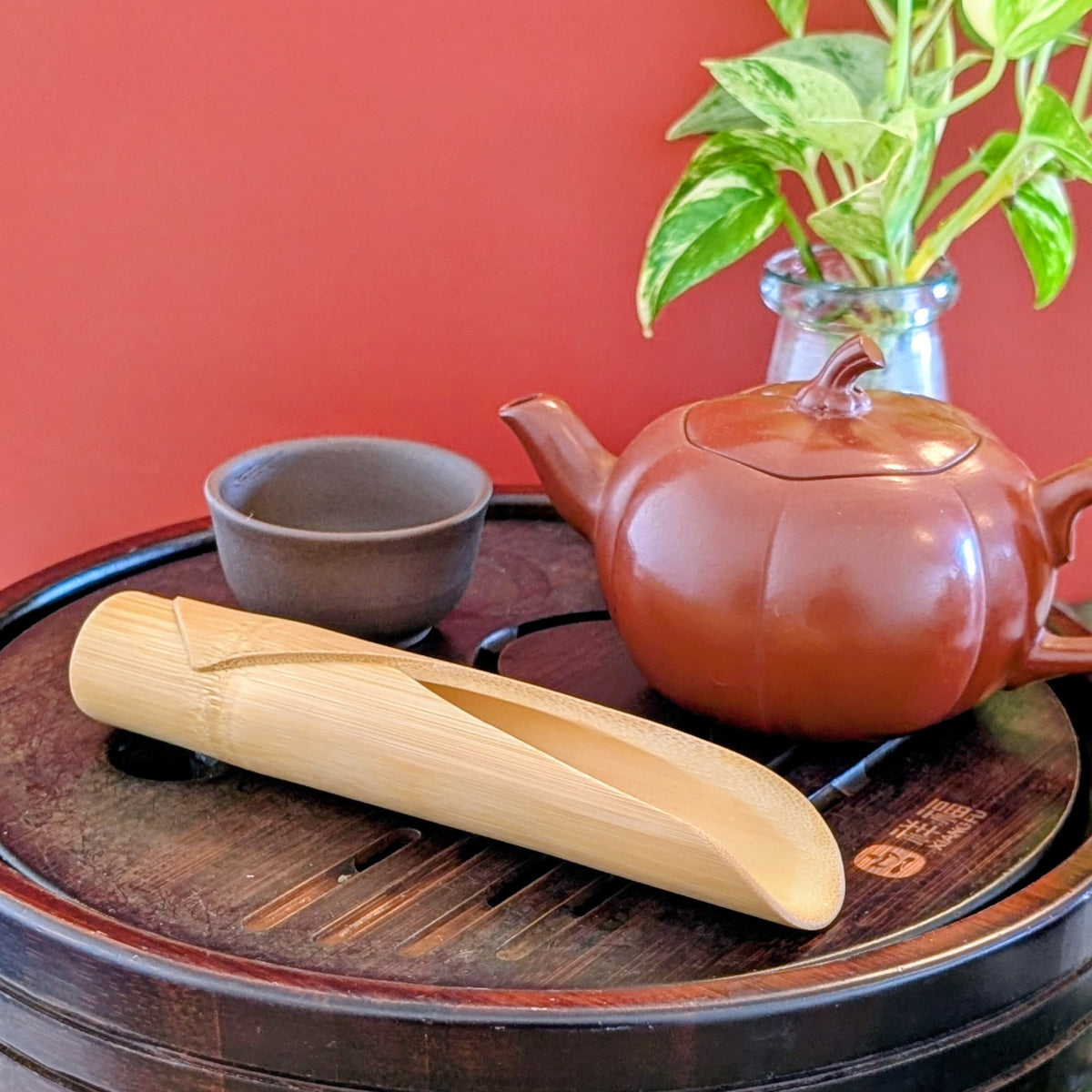 Bamboo scoop on a wooden tea tray with brown clay cup and red clay pot against a red background.