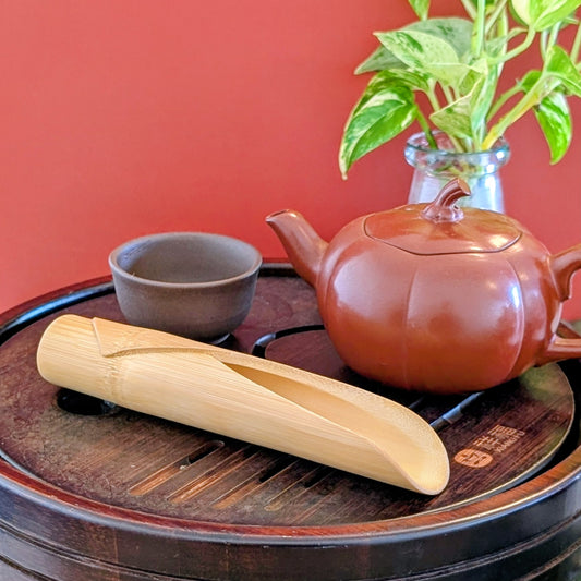Bamboo scoop on a wooden tea tray with brown clay cup and red clay pot against a red background.