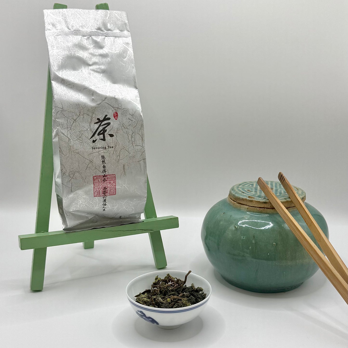 Tea packaging, green ceramic jar, bowl of tea leaves, and chopsticks on a white background