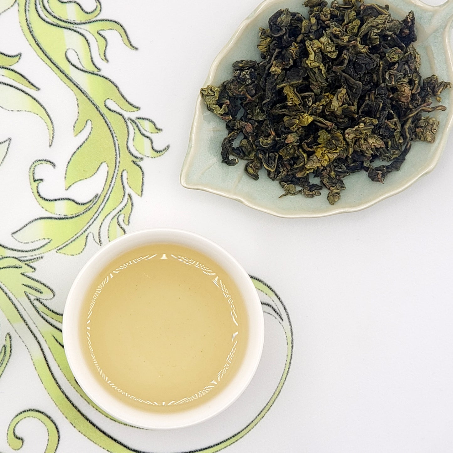 Dried tea leaves on a leaf-shaped plate with a cup of tea on a decorative mat.