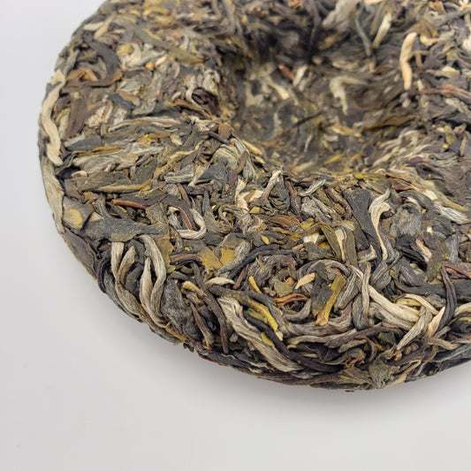 A close-up of a round compressed tea cake, showing the textured blend of dark and light leaves, on a plain white background.