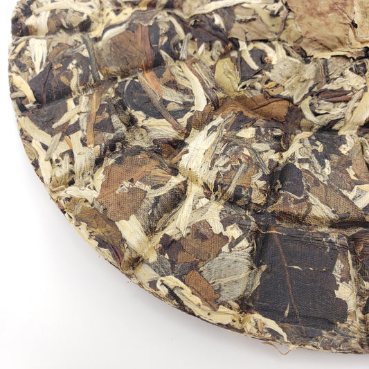 A close-up of a compressed tea cake, showing the textured, fibrous surface of the dried tea leaves and leaves, on a plain white background.