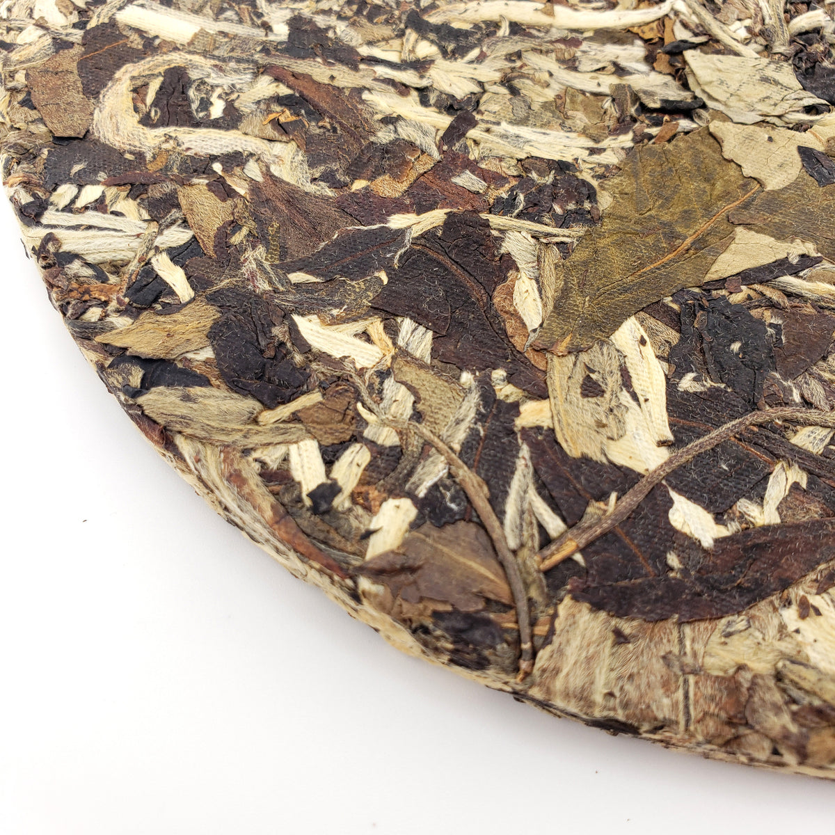 A close-up of a compressed cake of loose-leaf tea, showing mottled dark brown, tan, and off-white dried leaves and bark pieces, photographed on a white background.