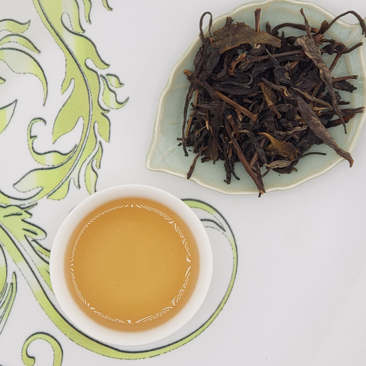 A cup of pale golden tea next to a dish of dried loose-leaf tea on a white surface with a green patterned design.
