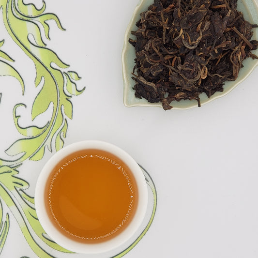Dried tea leaves on a white plate with a cup of brewed tea on a decorative white surface.