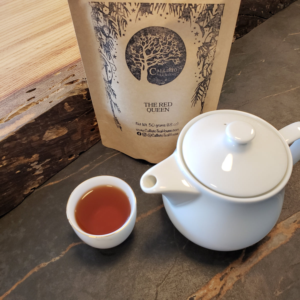 A white ceramic teapot with a lid, a small white cup filled with amber-red tea, and a kraft paper pouch labeled 'THE RED QUEEN' on a dark stone surface.