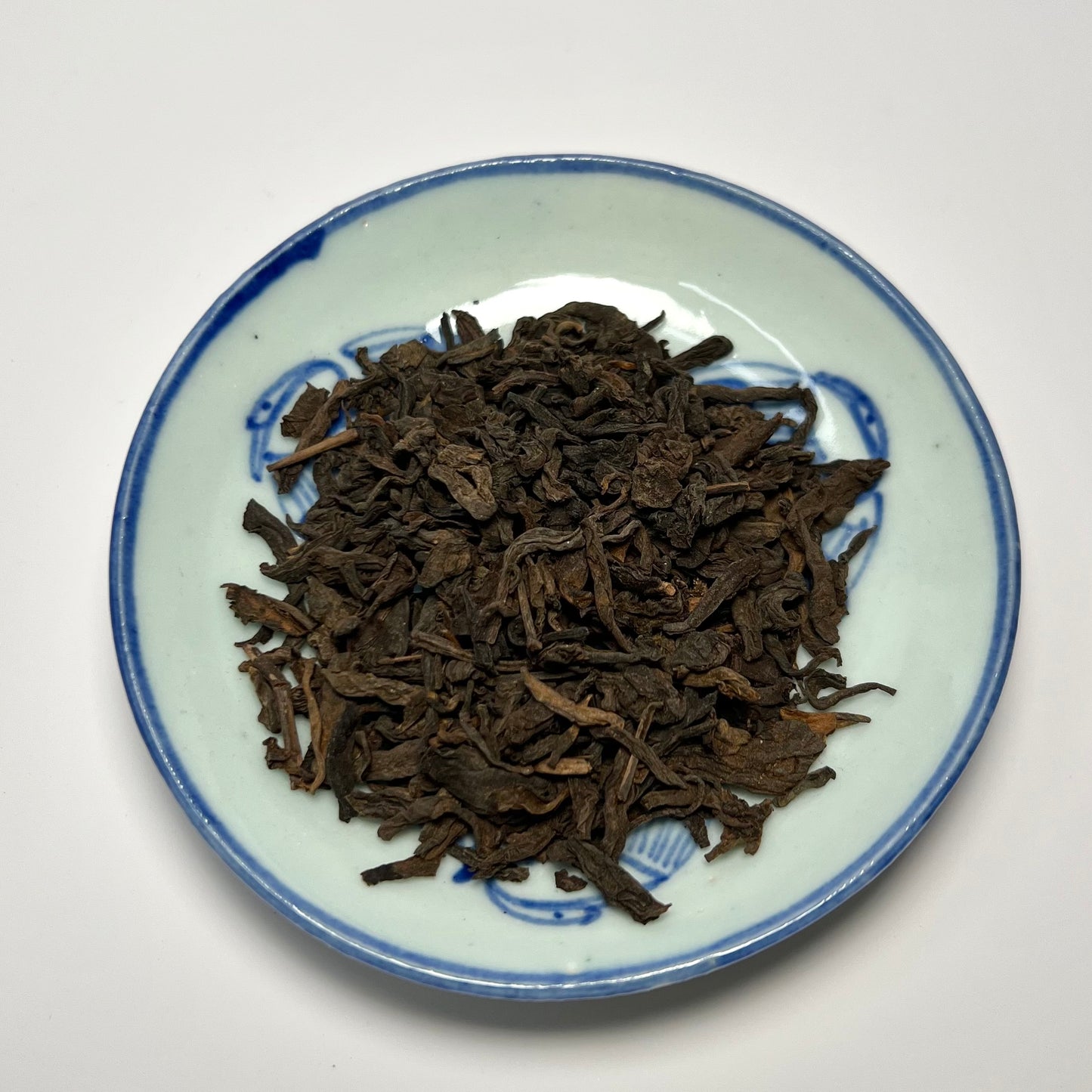 Dried tea leaves on a small ceramic plate with blue decorative rim against a white background