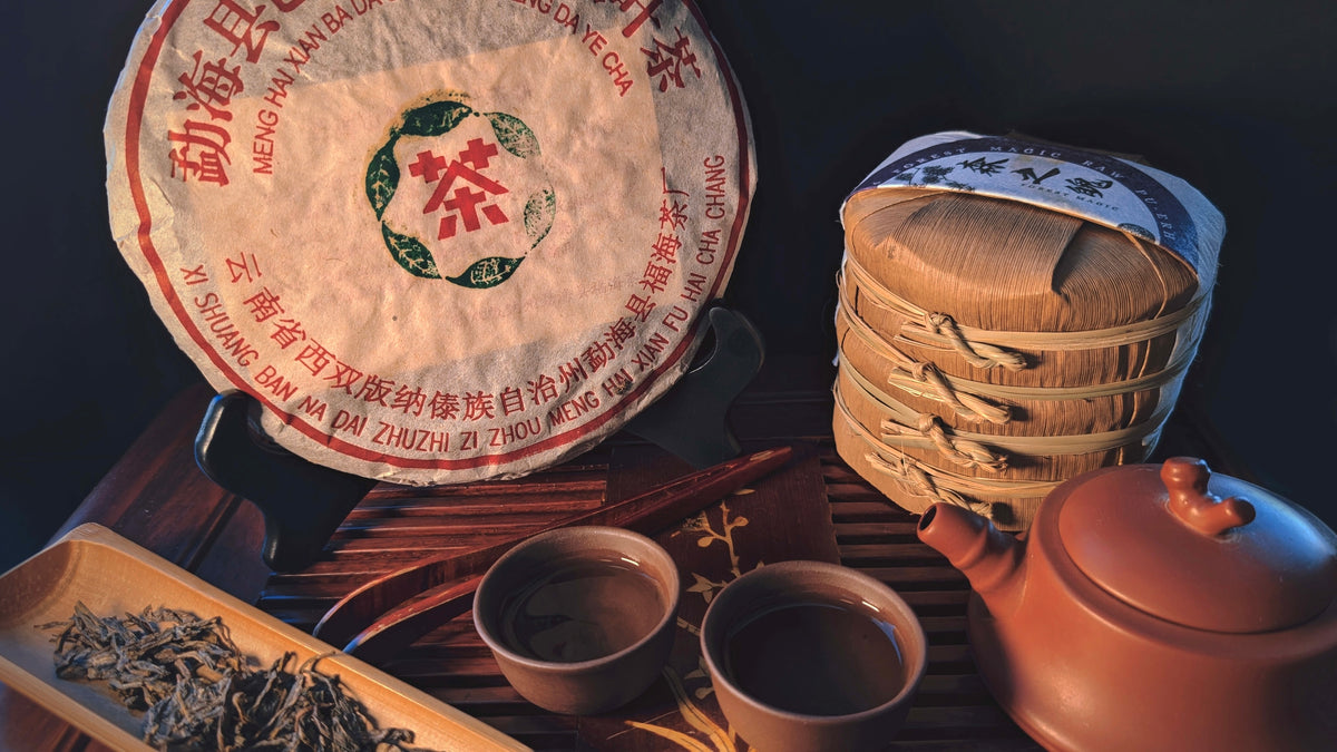 A round wrapped tea cake with Chinese characters and a green tea logo, displayed beside a stack of tea cakes wrapped in bamboo, two small tea cups, a terracotta teapot, and loose dried tea leaves on a dark wooden surface.