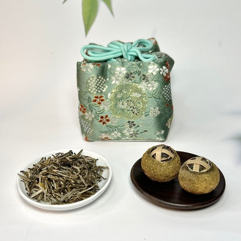 Tea leaves on a white plate, tea balls on a black plate, and a decorative tea bag with floral patterns.