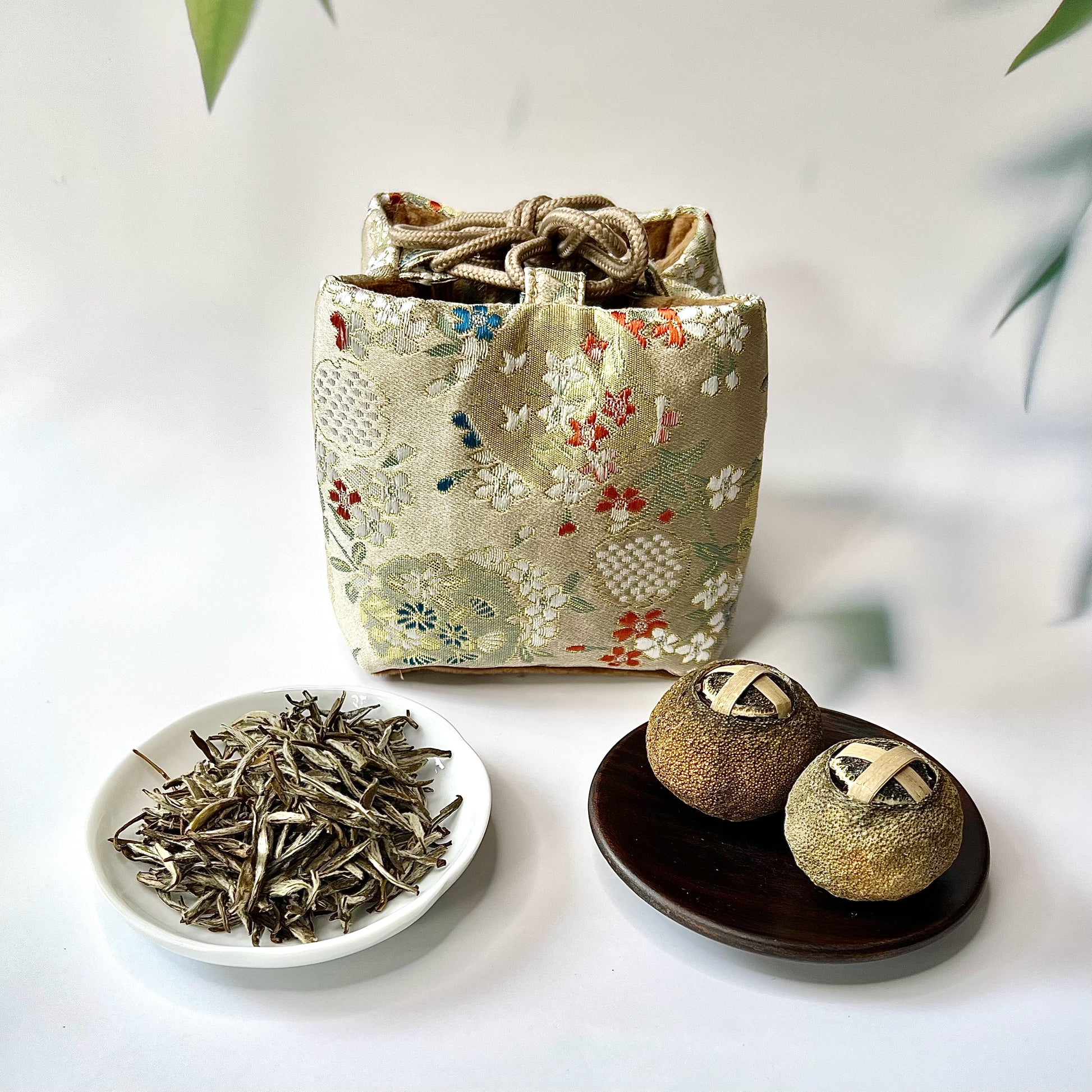 Fabric pouch with floral patterns, a white dish with tea leaves, and two round tea containers on a light background.