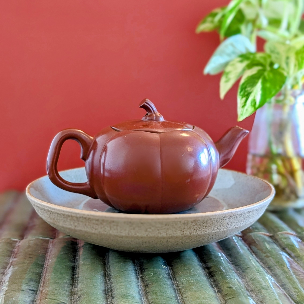 Red clay gongfu tea pot on shallow bowl or plate, grey speckled clay color, against a red background, on a green mat.