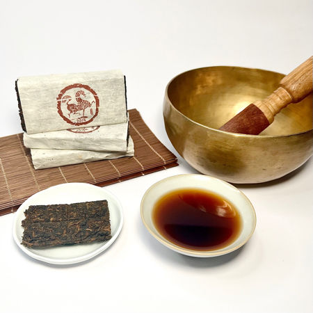 Tea packaging, a gold singing bowl, a small bowl of tea, and a leaf on a white background