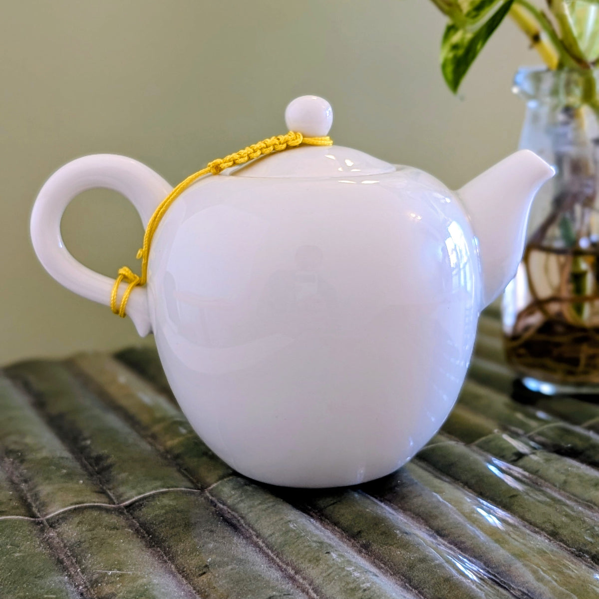 A glossy white ceramic teapot with a bright yellow braided cord handle, resting on a green bamboo mat with a blurred glass vase in the background.