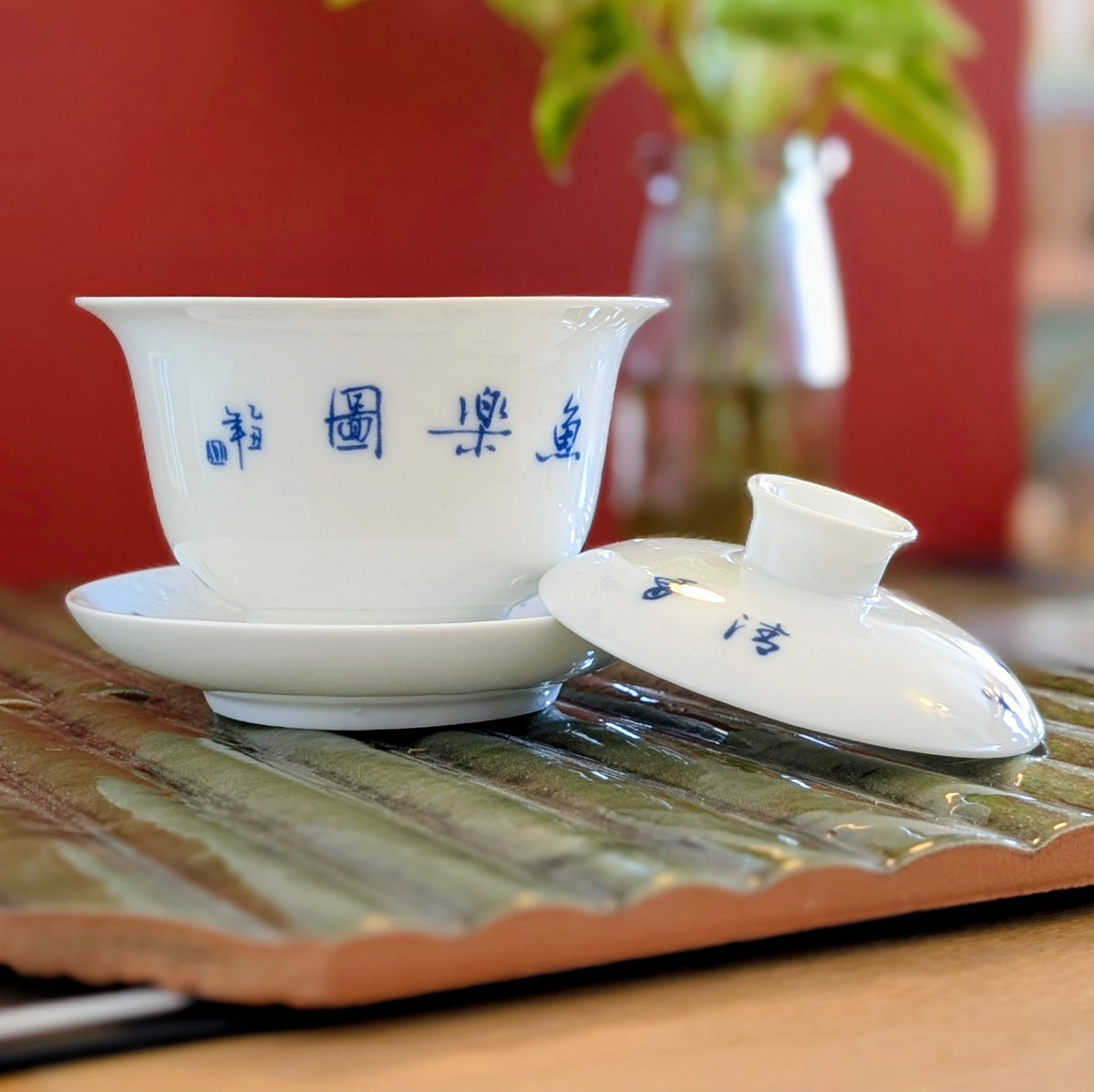 A white ceramic gaiwan set with a saucer and lid, decorated with blue Chinese calligraphy, displayed on a green bamboo tea tray with a warm red background.
