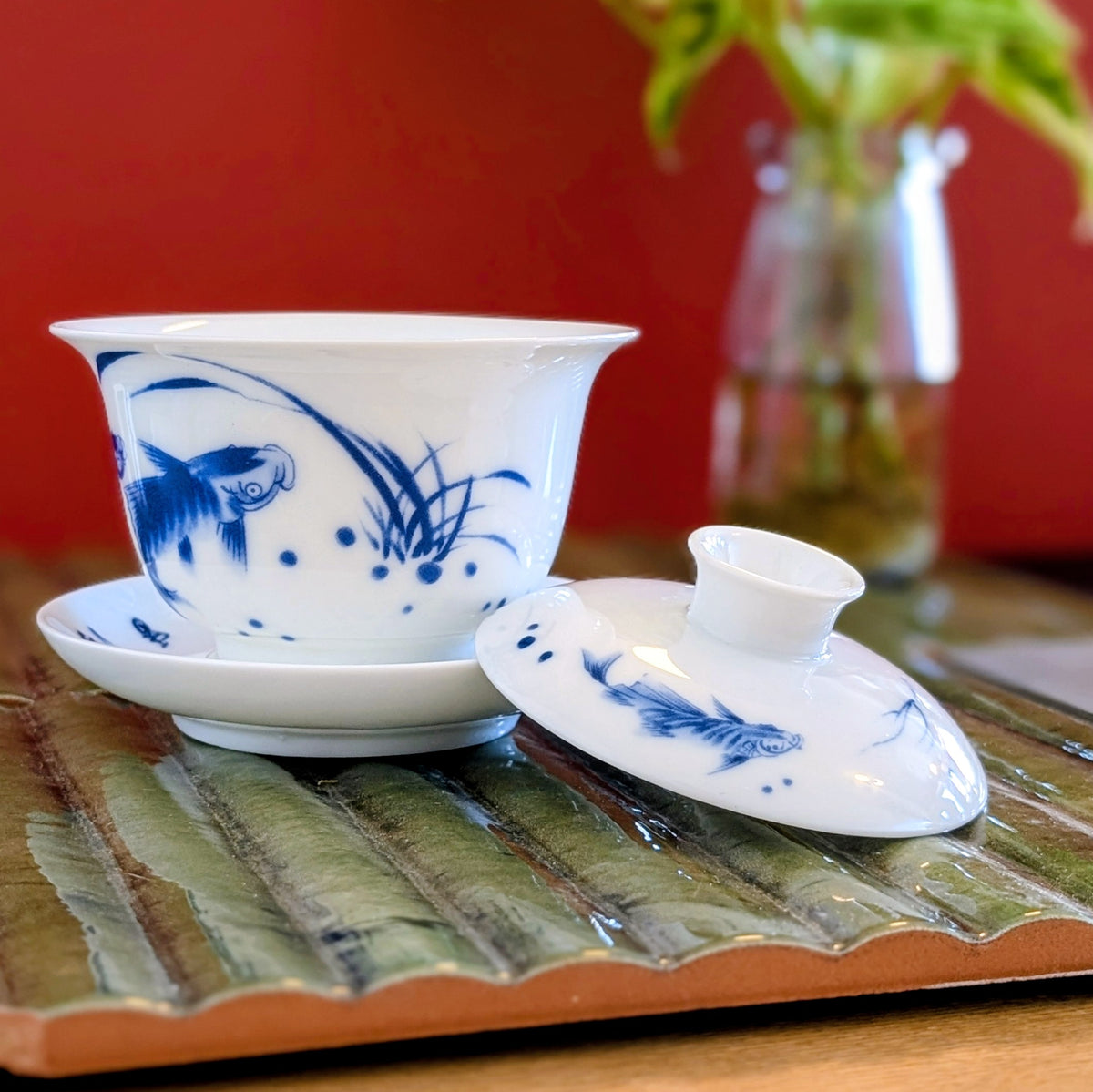 A white ceramic gaiwan tea set with blue fish and plant illustrations, shown with its saucer and lid on a decorative tray against a red background.