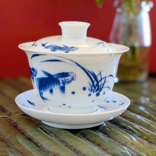 A white ceramic gaiwan tea set with blue hand-painted fish and lotus motifs, resting on a bamboo mat against a red background.