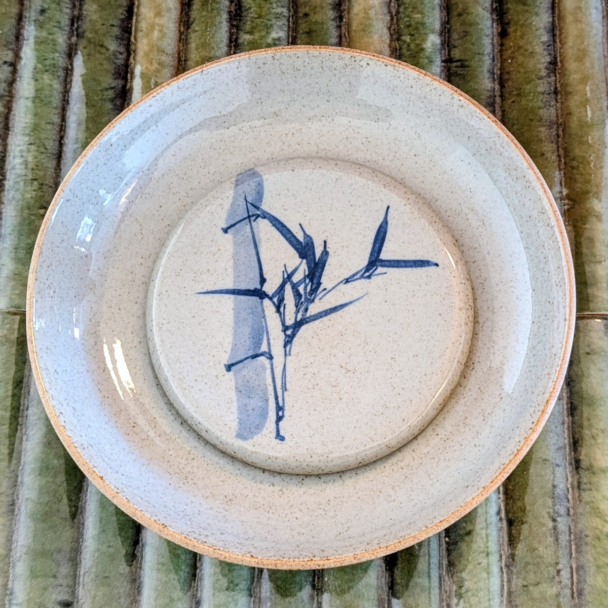 A round ceramic plate with a speckled beige glaze, featuring a blue bamboo illustration in the center and a natural unglazed clay rim, displayed on a green bamboo mat.