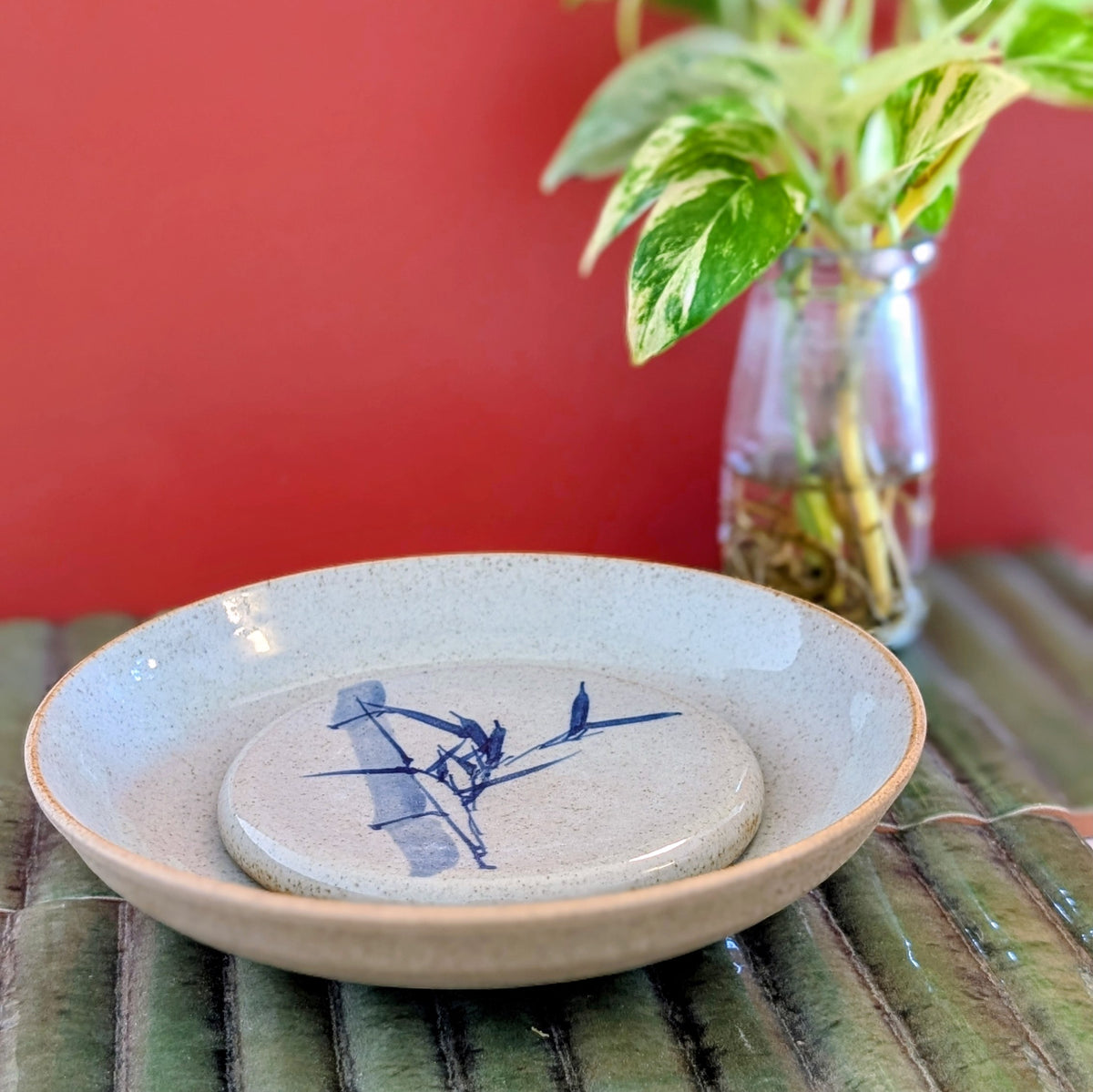 A ceramic round shallow plate in speckled beige with a blue painted bamboo design, set on a green bamboo mat with a red wall and a plant in the background.