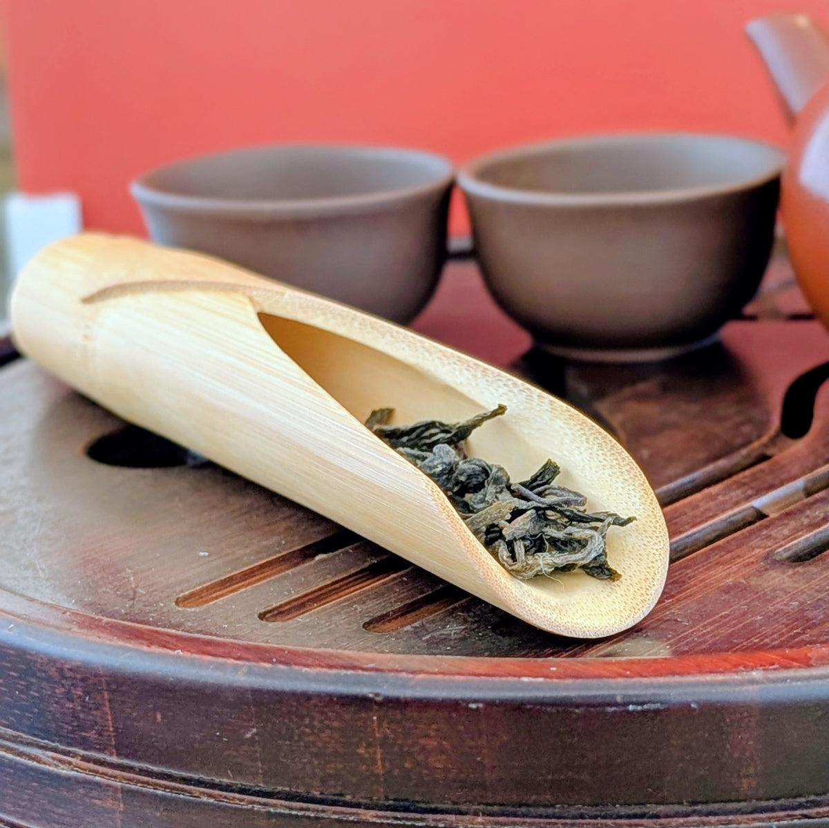Bamboo scoop with tea leaves on a wooden tray with clay tea cups against a red background.