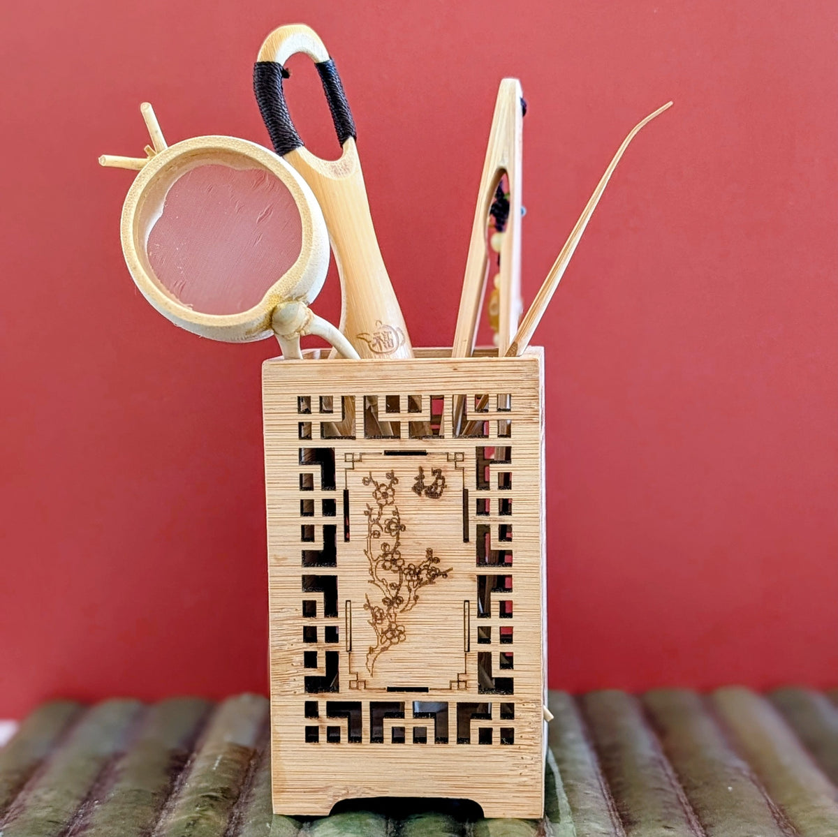 A light natural bamboo organizer holder with engraved floral design, holding tea tools like a strainer, scoop, tongs and pick, set against a solid red background.