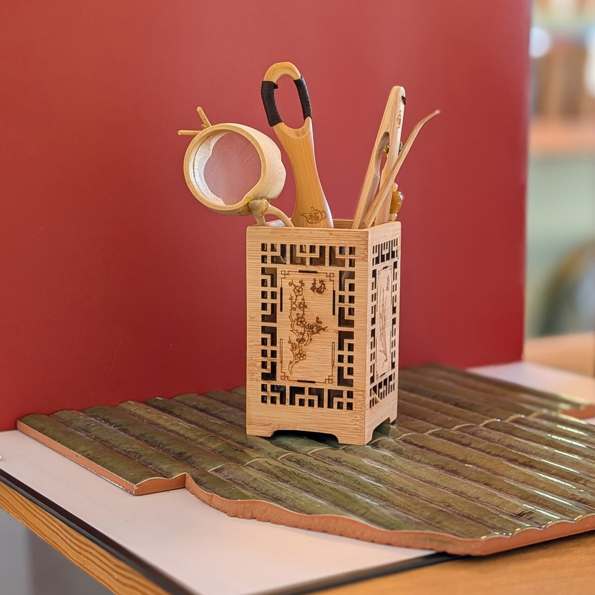 A light natural bamboo organizer holder with engraved floral design, holding tea tools like a strainer, scoop, tongs and pick, set against a solid red background.