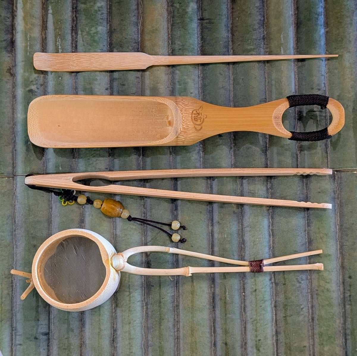 A bamboo and wood tea preparation set laid flat on a green textured background, including a scoop with black loop handle, tongs, a strainer, and small accessories.