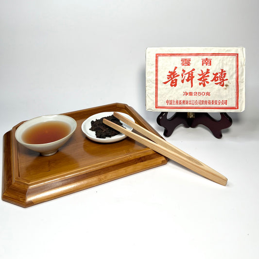 Tea set with a bowl, small plate, and tongs on a wooden tray with a tea bag in the background.