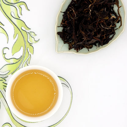 Dried pu-erh tea leaves on a leaf-shaped plate with a cup of brewed tea on a decorative white and green background.
