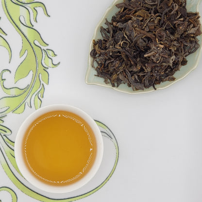 Dried tea leaves on a plate with a cup of brewed tea on a decorative mat.