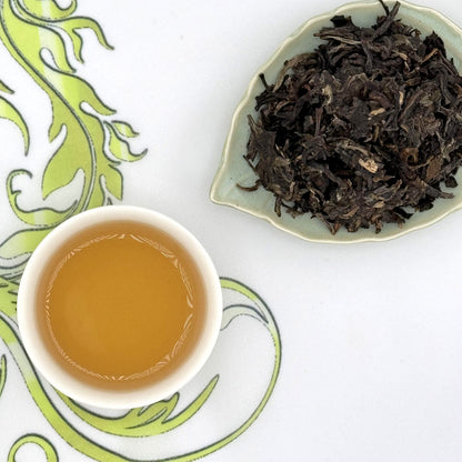 Tea cup with brewed tea and a dish of tea leaves on a decorative white and green surface.