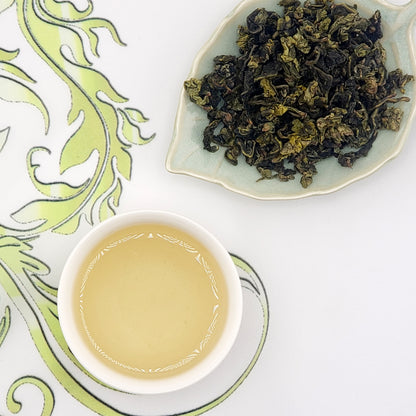 Dried tea leaves on a leaf-shaped plate with a cup of tea on a decorative mat.