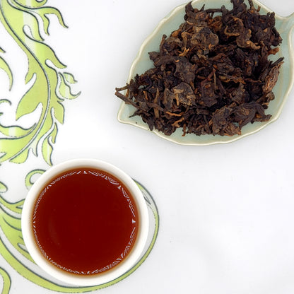Dried tea leaves on a leaf-shaped plate with a cup of brewed tea on a decorative mat.