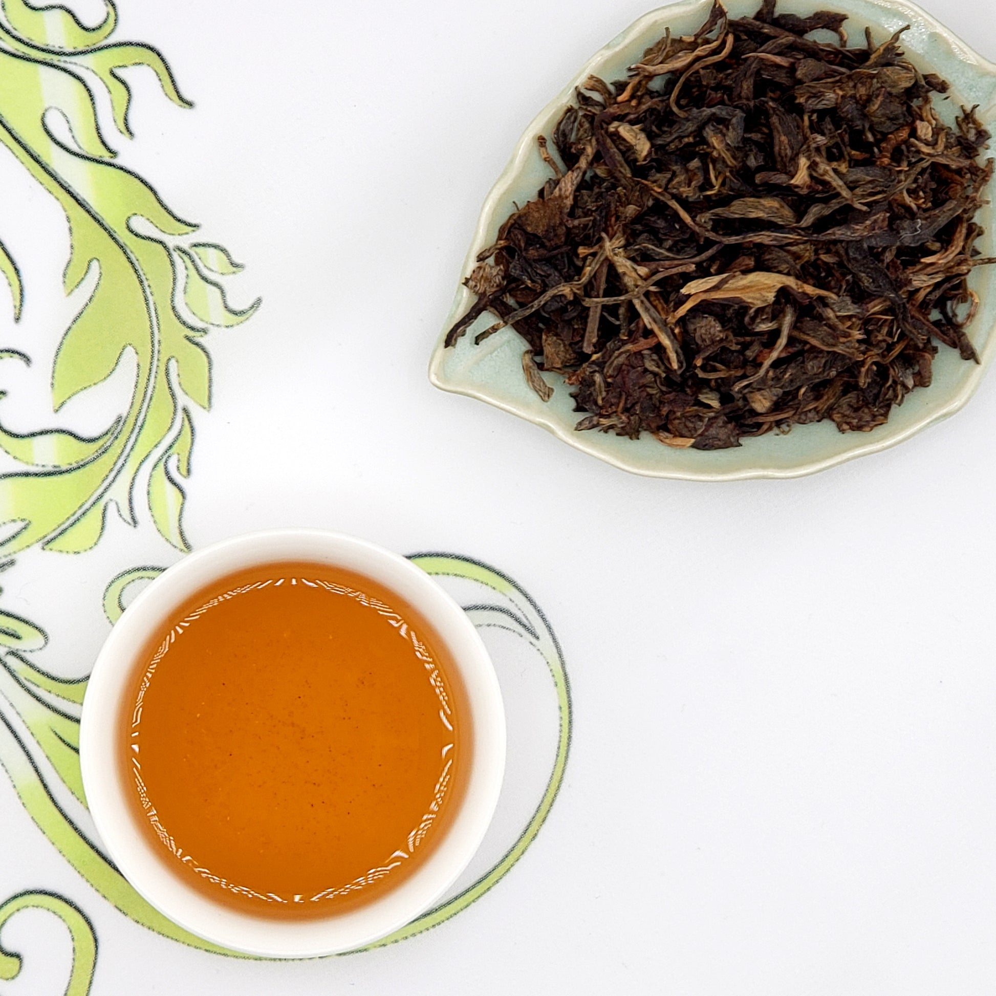 Dried tea leaves on a leaf-shaped dish with a cup of brewed tea on a decorative tablecloth.