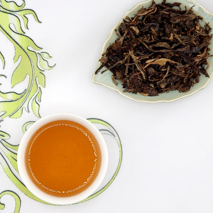 Dried tea leaves on a leaf-shaped dish with a cup of brewed tea on a decorative tablecloth.