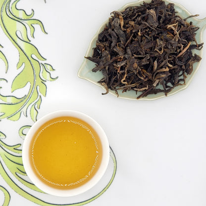 Dry tea leaves on a leaf-shaped plate with a cup of brewed tea on a decorative white and green background.