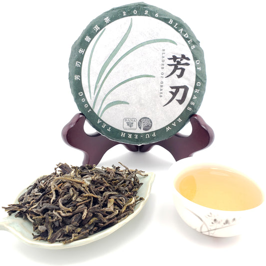 A round cake of Pu-erh tea displayed on a stand, with loose dried tea leaves in a small dish in front and a cup of pale tea on the right, all on a white background.