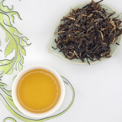 Dry tea leaves on a plate with a cup of brewed tea on a decorative white and green background.