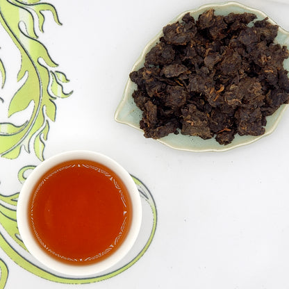 Dried tea leaves on a green leaf-shaped plate with a cup of brewed tea on a white background.
