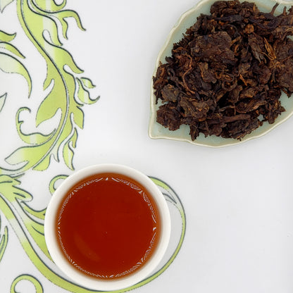 Dried tea leaves on a plate and brewed tea in a cup on a decorative tablecloth.