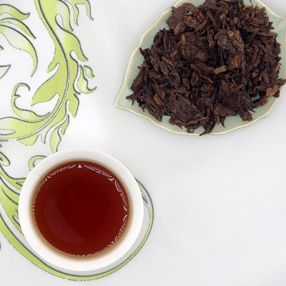 Dried tea leaves on a plate with a cup of brewed tea on a decorative mat.