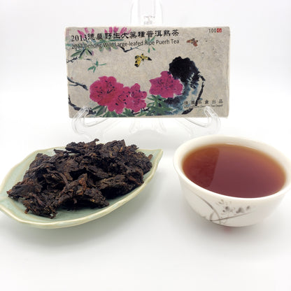 Pressed brick of Pu-erh tea with decorative wrapper, plate of dry tea leaves broken from cake, and a cup of brewed tea, dark amber in color.