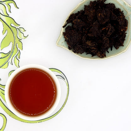 Dried tea leaves on a leaf-shaped plate with a cup of brewed tea, rich dark amber color, on a decorative mat.