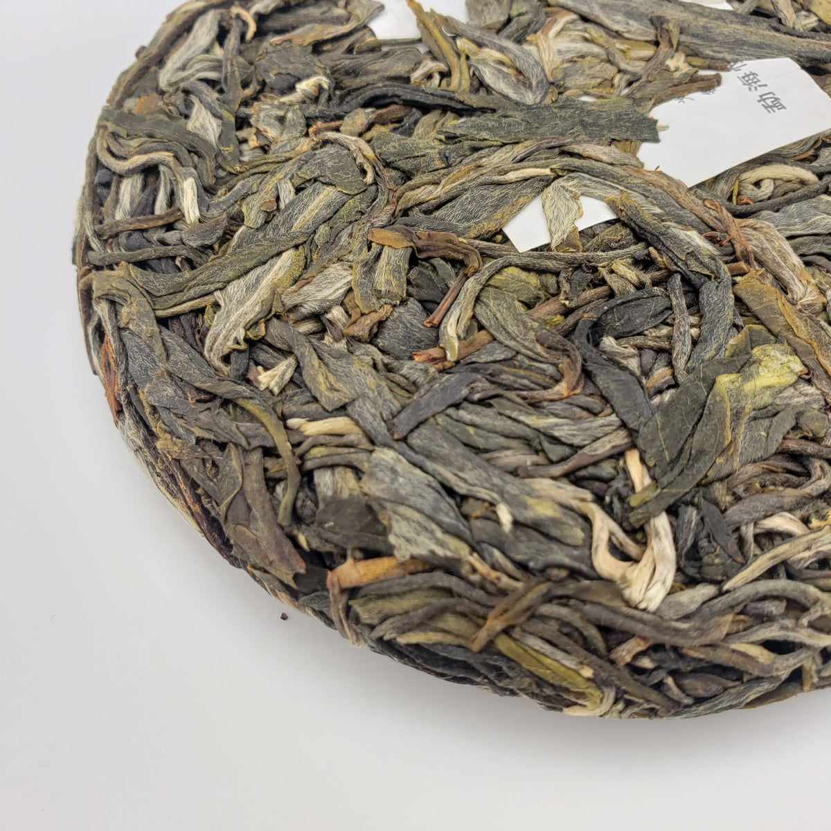A close-up of a round compressed cake of loose-leaf tea, showing tightly packed dried leaves in muted green, brown, and off-white tones, with a small white paper tag visible on the upper right.