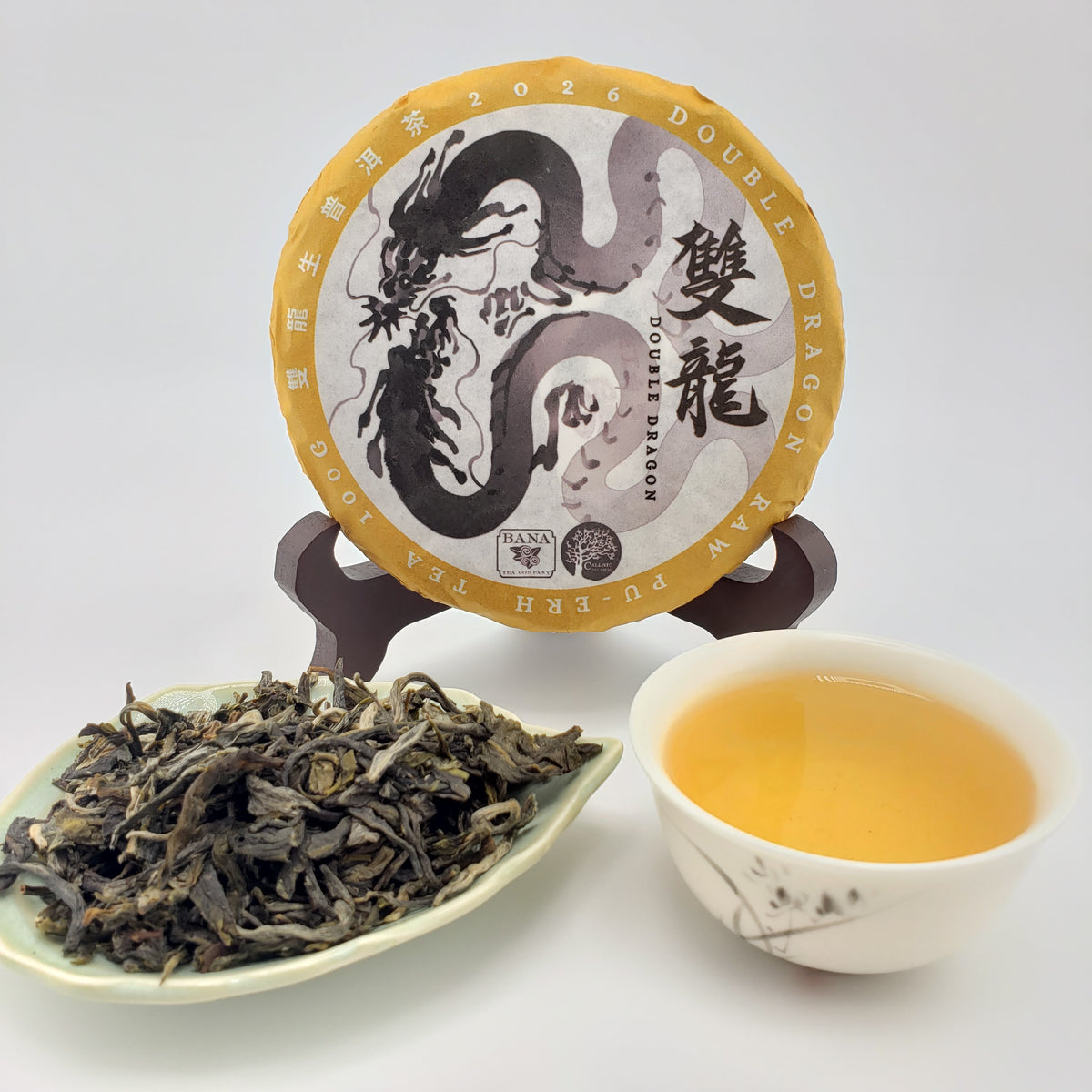 A round wrapped tea cake displayed on a stand, with a small dish of loose dried tea leaves and a cup of brewed yellow tea on a white background. The tea label shows a dragon illustration and Chinese characters, including 'DOUBLE DRAGON'.