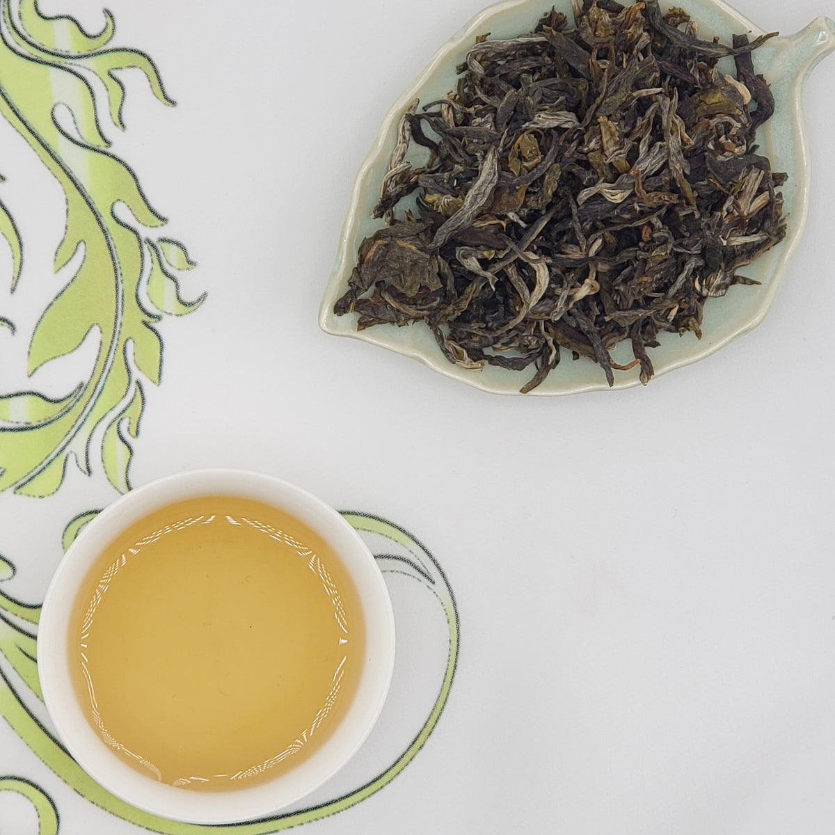 A cup of pale golden tea on a white saucer next to a leaf-shaped dish filled with dried loose-leaf tea, arranged on a white surface with a green patterned cloth.