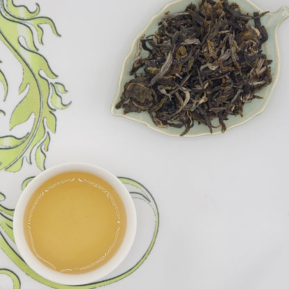 A cup of pale golden tea on a white saucer next to a leaf-shaped dish filled with dried loose-leaf tea, arranged on a white surface with a green patterned cloth.