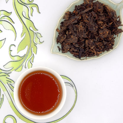 Dry tea leaves on a leaf-shaped plate with a cup of brewed tea on a decorative surface.