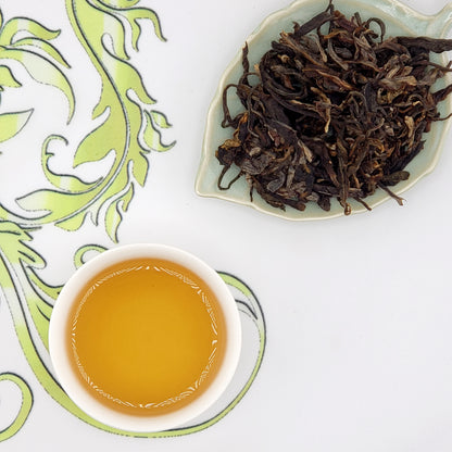 Dried tea leaves on a leaf-shaped dish with a cup of brewed tea on a decorative mat.